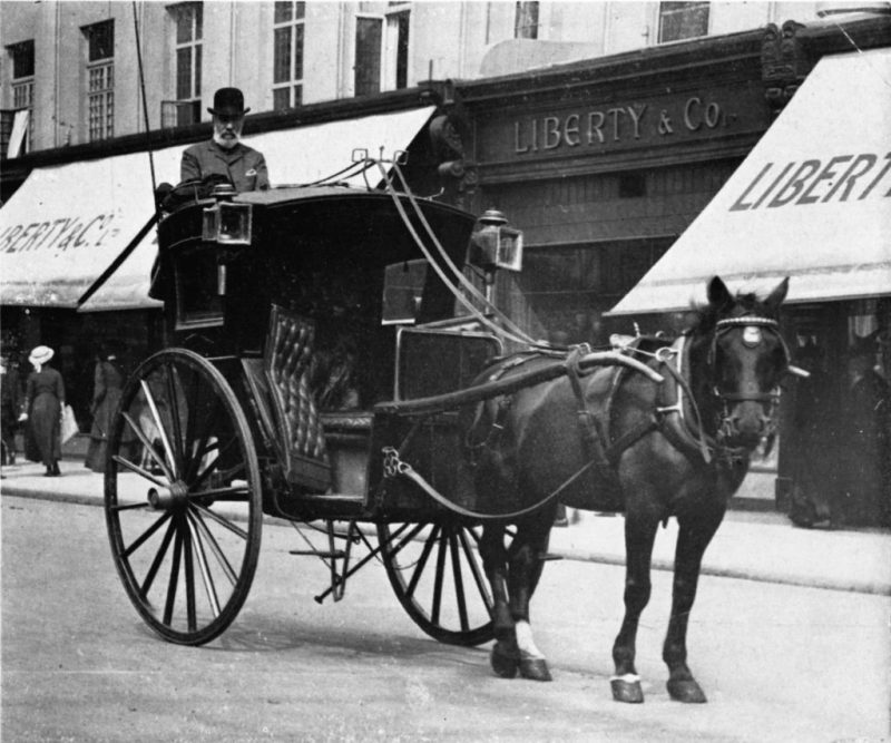 A black and white photo of an empty hackney coach in the middle of a street.