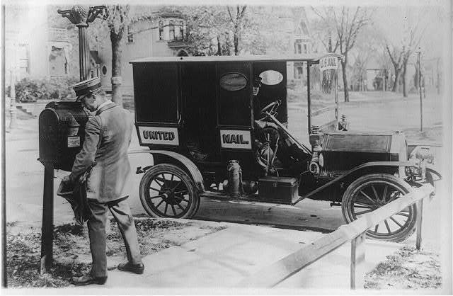 A black and white photo of a man standing at a mail post and a United Mail car parked on the side.