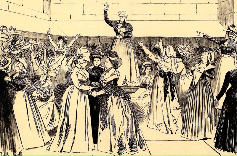 An all-women gathering with a woman on a stage in the middle of the room raising her right hand.