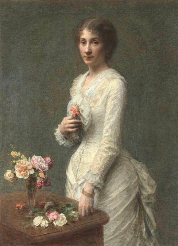 A lady holding a pink flower to her chest standing beside a table with a vase full of flowers.